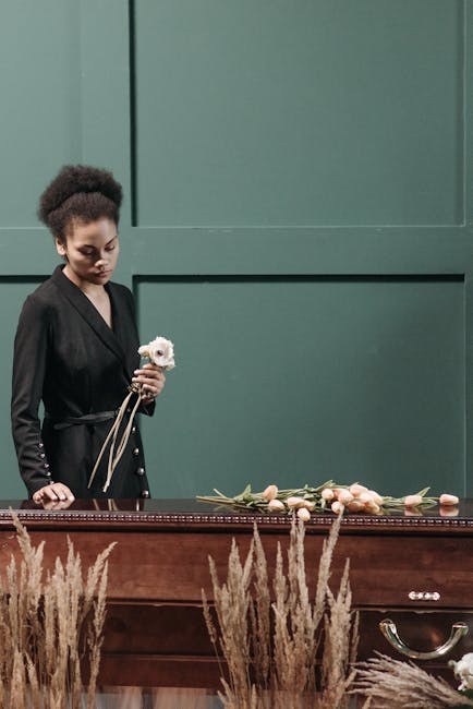 A young woman with curly dark hair wearing a black blazer stands behind a wooden casket, holding a single white flower in her right hand. In front of her, a floral arrangement of pale peach and cream