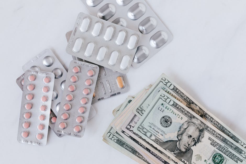 Close-up of multiple blister packs of pink, orange, and white pills arranged on a white surface, alongside a small stack of US twenty-dollar bills. The blister packs contain capsules and tablets, some
