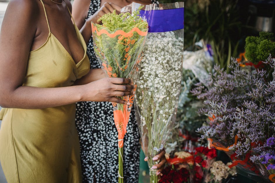 A woman wearing a yellow sleeveless dress is holding a bouquet of mixed flowers wrapped in clear and orange floral paper, with visible green foliage and small white blooms. Behind her, another person