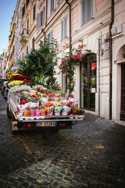A delivery truck parked outside a beige, multi-story building with classic architectural details and window shutters, loaded with a large array of fresh flowers arranged in various vases, bouquets, an