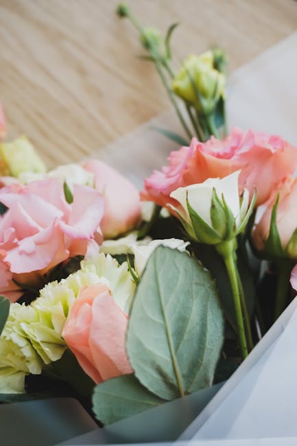 A close-up view of a bouquet featuring pale pink and cream Lisianthus flowers with ruffled petals, complemented by fresh green leaves and stems. The bouquet is partially wrapped in white tissue paper