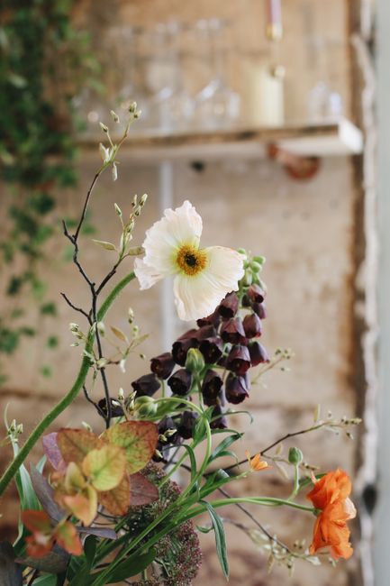A close-up of a floral arrangement featuring a white poppy with delicate, slightly ruffled petals and a dark center, alongside dark purple bell-shaped flowers and orange blooms, all with fresh green f