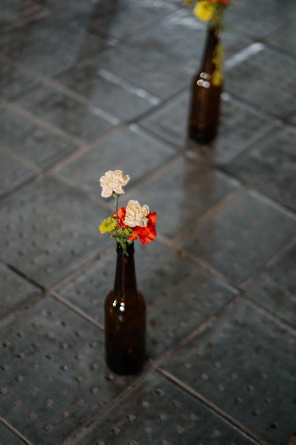 A slender, dark amber glass bottle holds a small floral arrangement featuring white carnations, a single red carnation, and some yellow-green foliage, placed on a textured stone pavement illuminated b