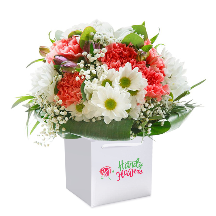 Expertly designed for moments of sincere celebration, this lush bouquet sits upright in a sharp, clean white square gift bag, photographed on a luminous white background that amplifies every detail. The arrangement bursts forth with soft coral-pink and delicate peach carnations, each petal frilled and richly textured, contrasted beautifully with the joyful presence of white daisies, their bold yellow-green eyes gleaming with optimism. Tiny clusters of white baby's breath are woven throughout, introducing an airy gentleness and echoing the lightness often felt strolling through Shacklewell's open spaces. Pops of unopened buds, tinged in subtle purple, rest quietly on the left, while sleek, broad green leaves complete the display, creating a natural, almost sculptural collar around the floral heart. The sturdy white ribbon handles above the bag hint at easy delivery to any doorstep along Shacklewell Lane or a welcoming café off Amhurst Road. Arranged in a balanced, rounded fashion, the bouquet expresses effortless abundance - an embodiment of freshness and care, reflective of regional expertise. The whole presentation conveys a sense of occasion: whether for a birthday, an anniversary, or simply to lift a loved one's spirits, this bouquet makes sending beauty simpler. Every stem is precisely placed, a testament to a local florist's skilled touch, ensuring that every recipient in Shacklewell receives both a gift and a gesture of heartfelt connection.